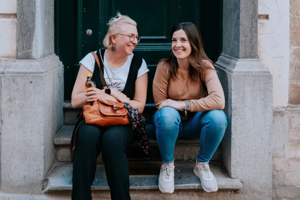 Deux jeunes artistes rigolent ensemble, assises sur devant une porte de maison.