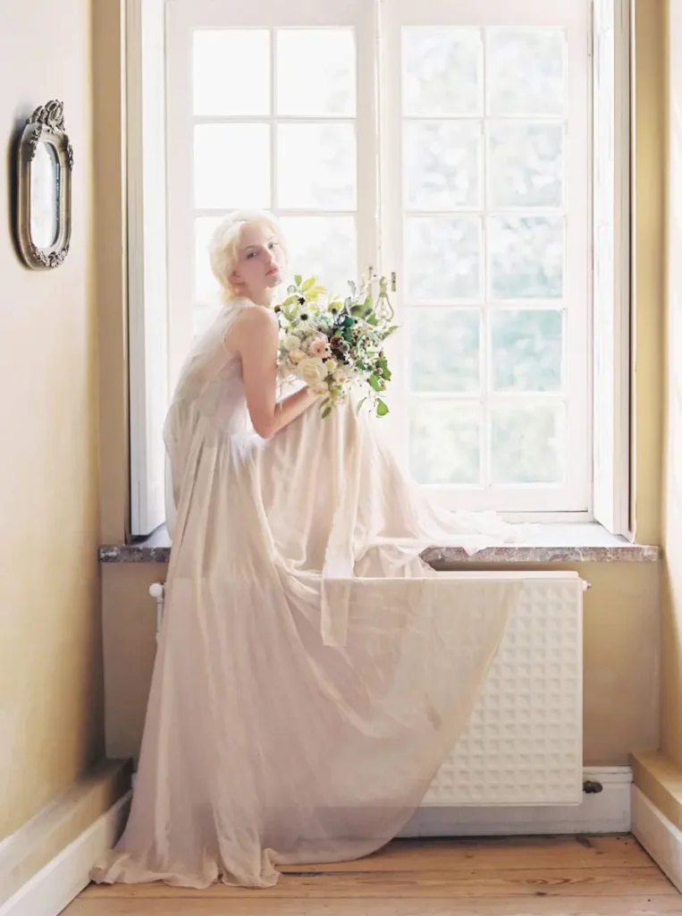 Portrait argentique d'une femme en longue robe assise sur bord de fenêtre avec un bouquet de fleurs.